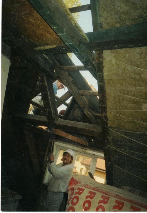 Pracownik w czapce z papierosem w ustach, w trakcie prac związanych z izolacją poddasza, widoczne drewniane elementy konstrukcyjne dachu i częściowo ułożona wełna mineralna, pakunki wełny...